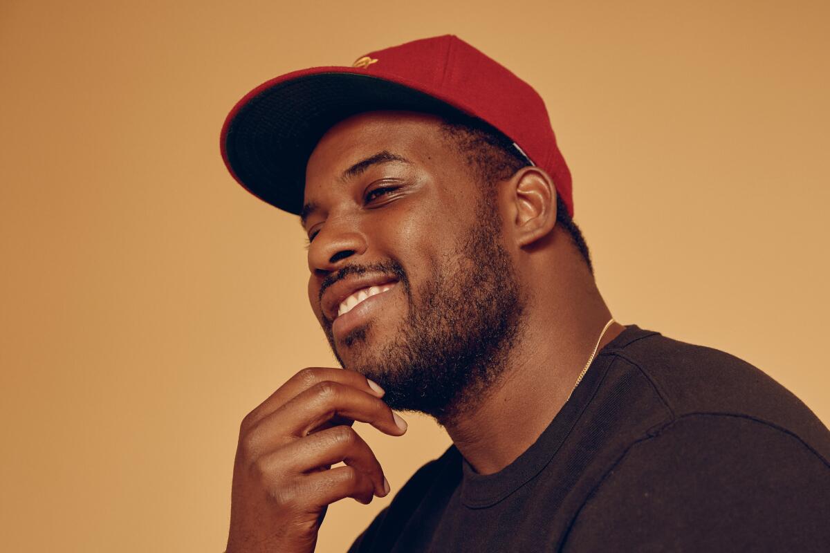 Lionel Boyce tilts his head and puts a hand to his chin. He wears a red baseball cap and black T-shirt.