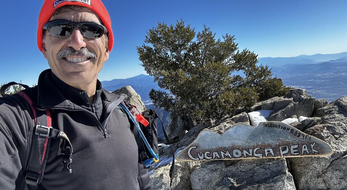 A smiling hiker taking a selfie near a sign reading "Cucamonga Peak."
