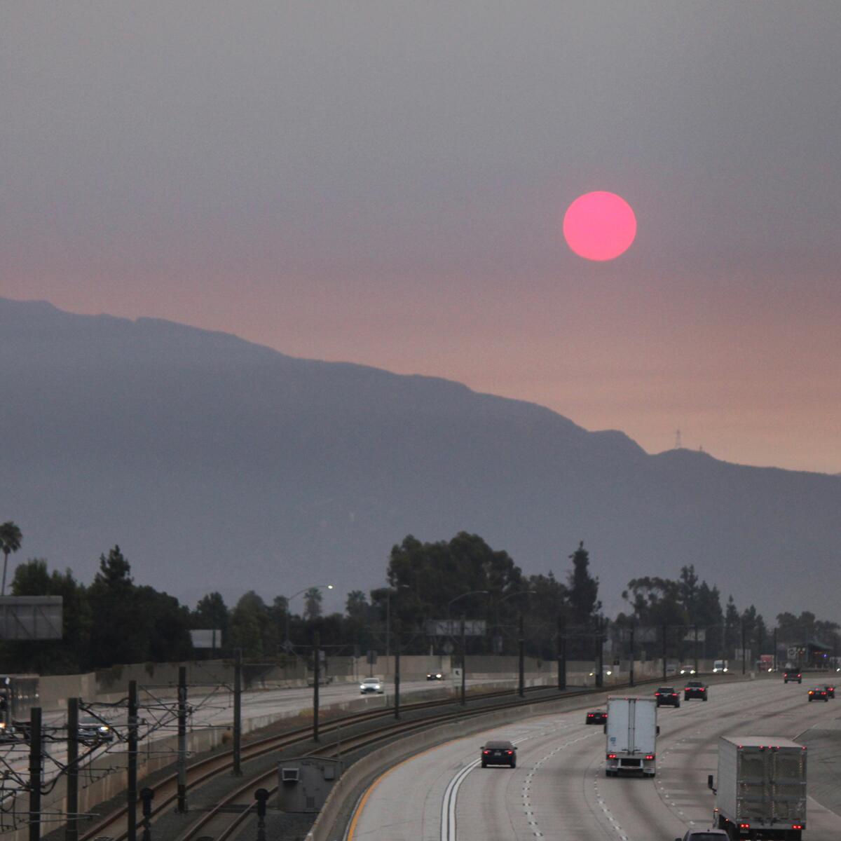 The setting sun, above a freeway and mountains, is pink amid dense wildfire smoke. 