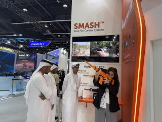 Men in white robes and headdresses watch as a woman takes aim with an orange rifle in a display area