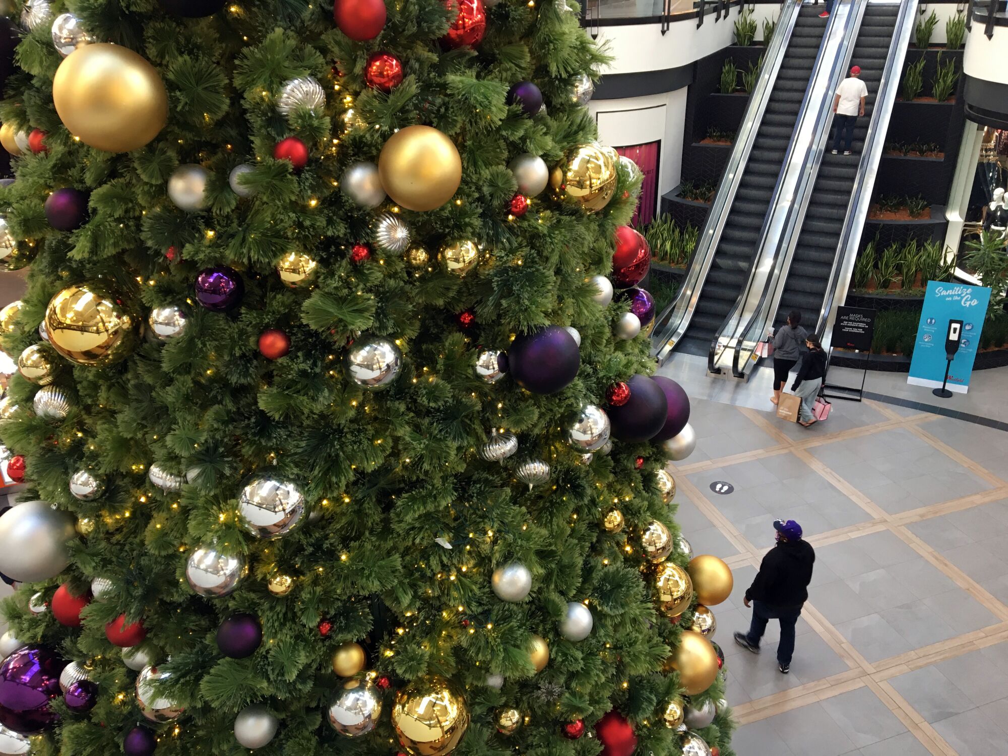 A huge Christmas tree dominates a mall courtyard as a man walks by and a few others ride an escalator. 