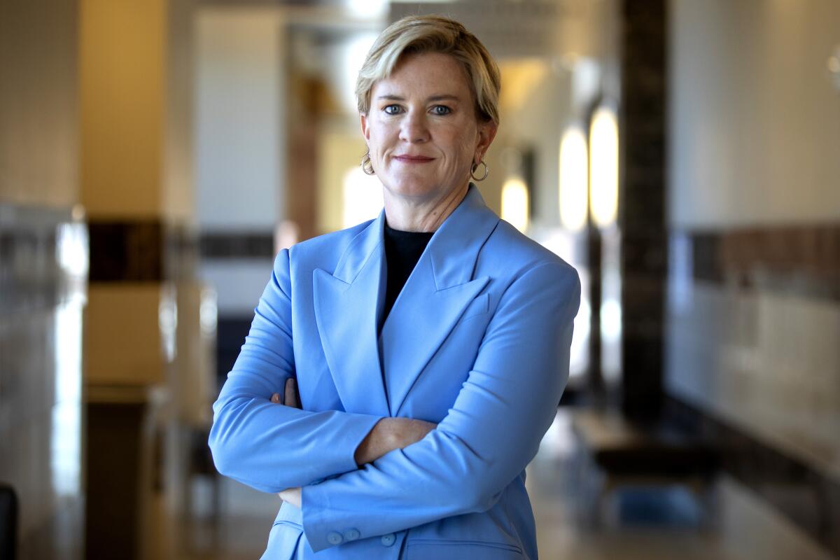 A smiling woman wearing a light blue blazer stands in a hallway, arms crossed