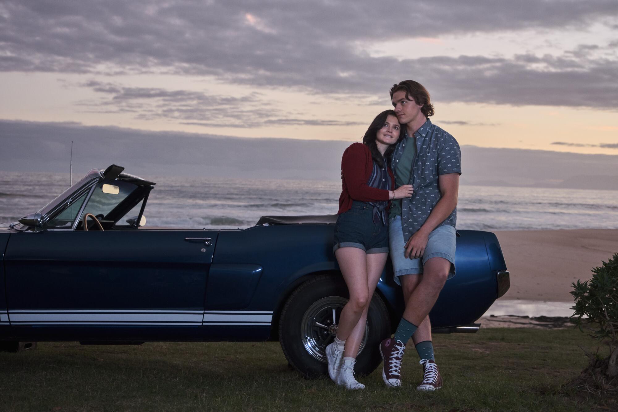 A man and a woman lean into each other as they lean onto a convertible car at the beach