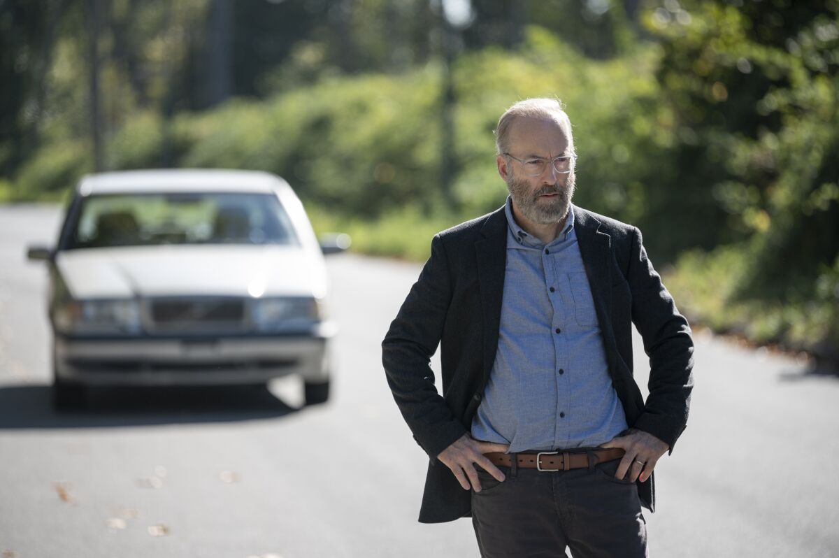 A man, wearing a blue shirt and dark blazer, stands on a road with his hands on his hips.