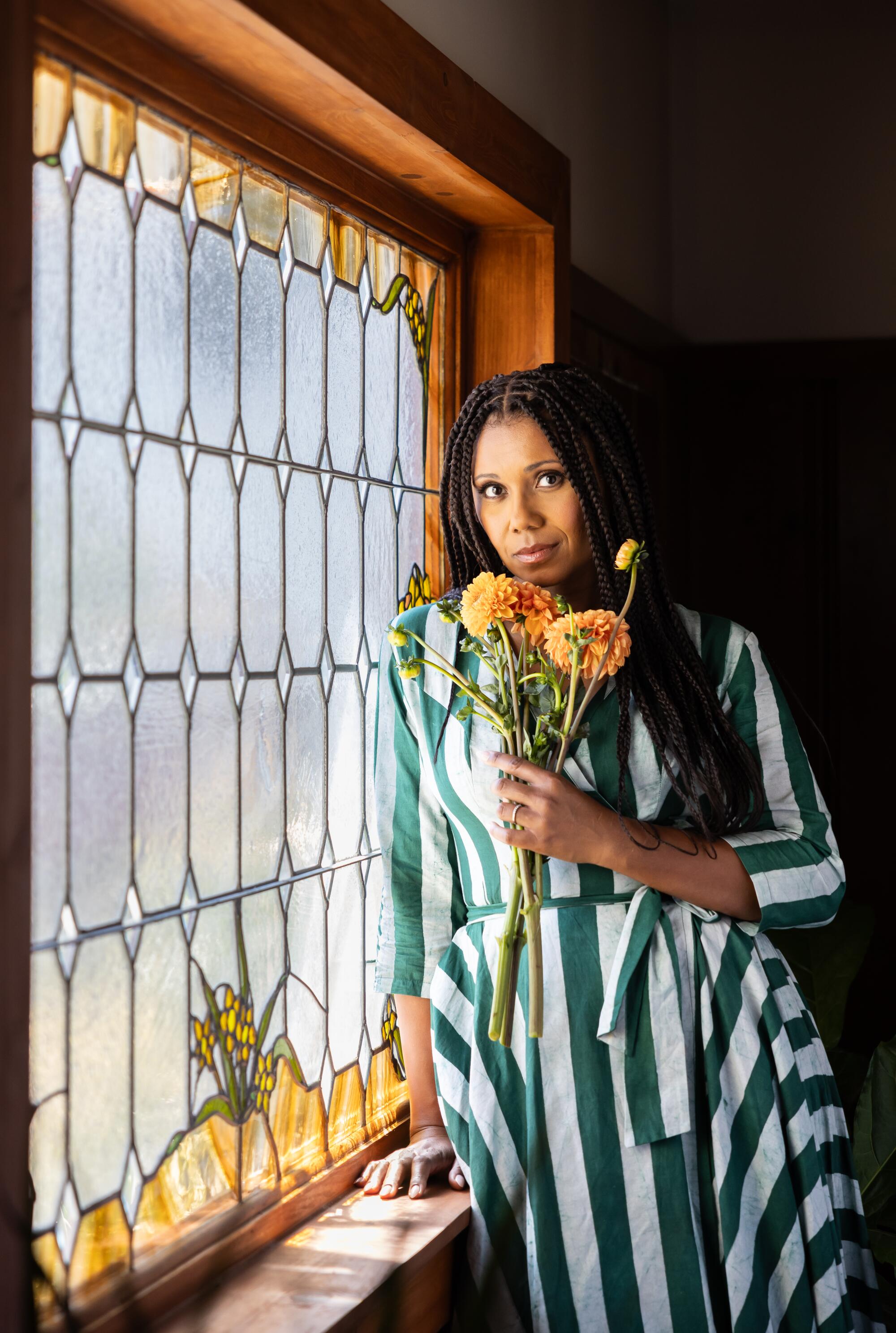 A woman holding flowers leans against the frame of a stained-glass window.