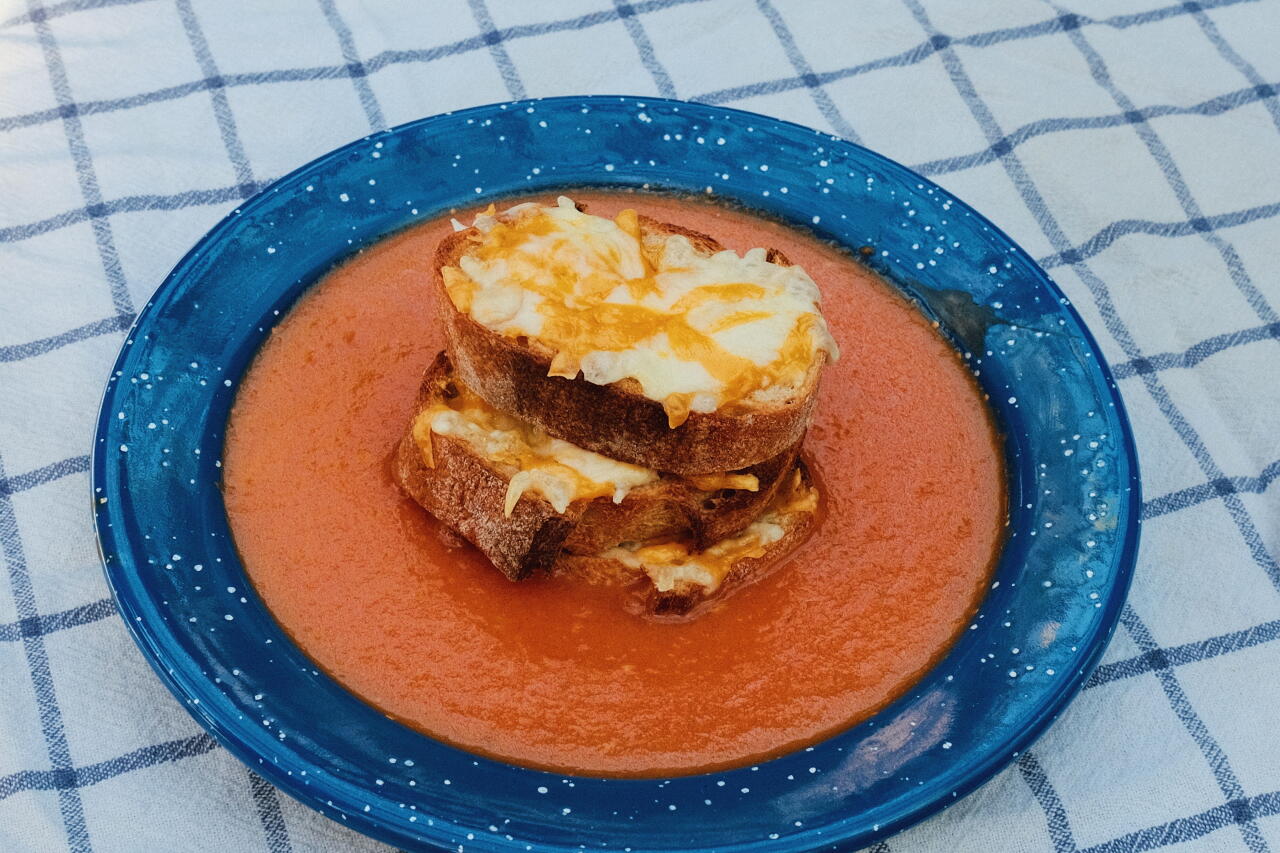 A blue enamel bowl of tomato soup on white-and-blue checkered cloth, with a stack of cheese-topped crostini