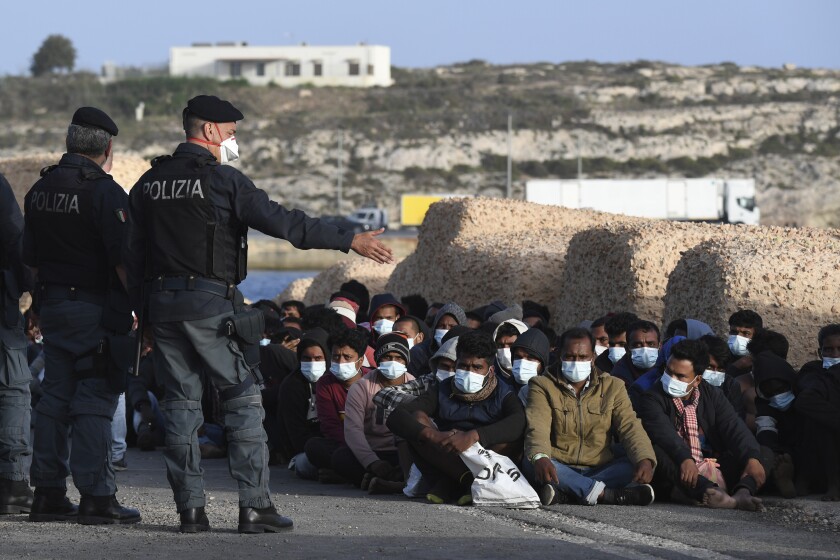 Migrants wearing face masks sit at a pier as Italian police officers stand by, on the Sicilian island of Lampedusa