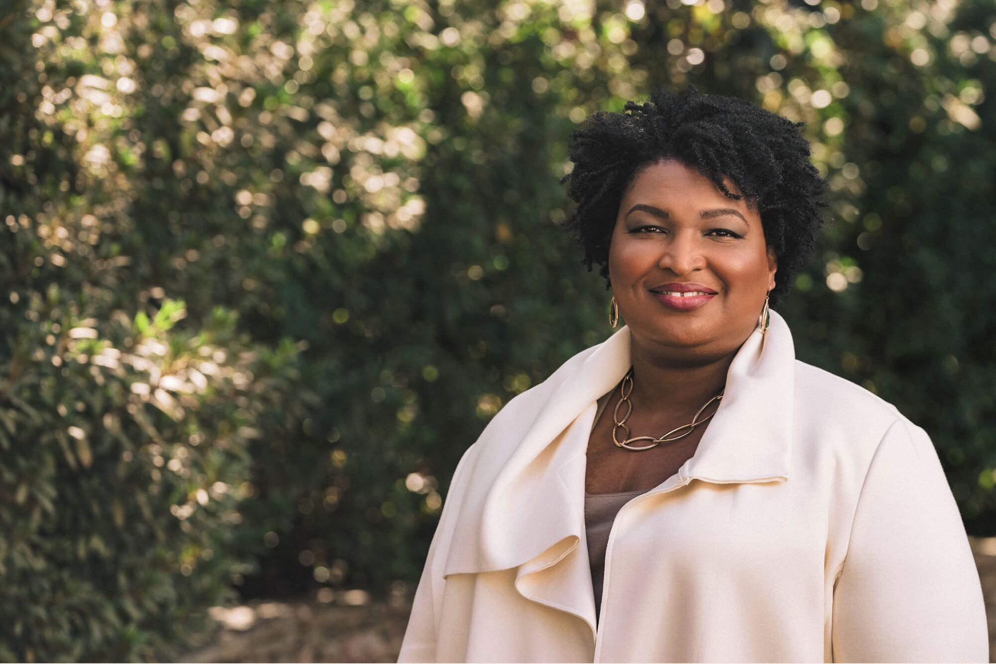 Stacey Abrams  smiles while standing in front of a group of trees and bushes.