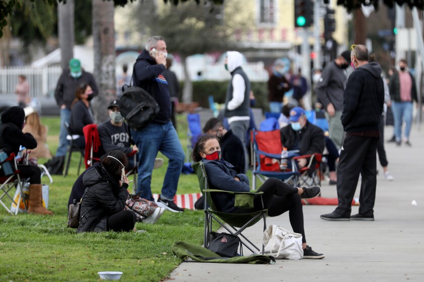 People wearing masks sit in folding chairs on the sidewalk and on the grass.