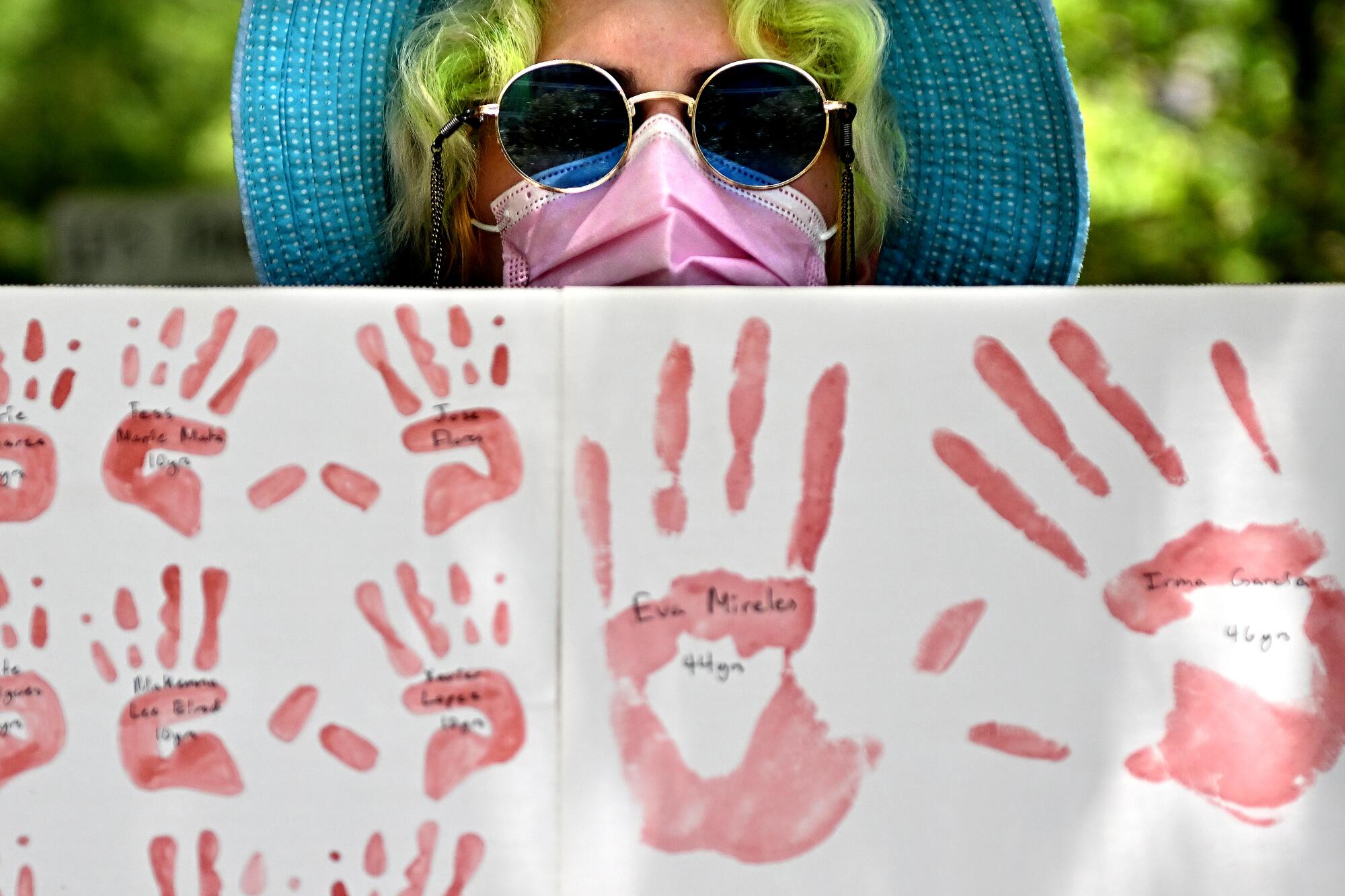 A person wearing a hat and sunglasses holds a posters with red handprints