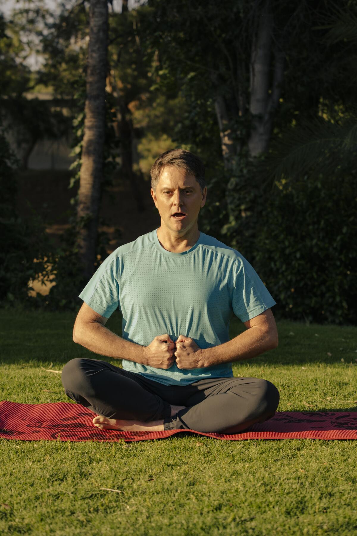 A man sits crosslegged on a yoga mat, his fists flat against his belly and his mouth open as he yells "Ha!"