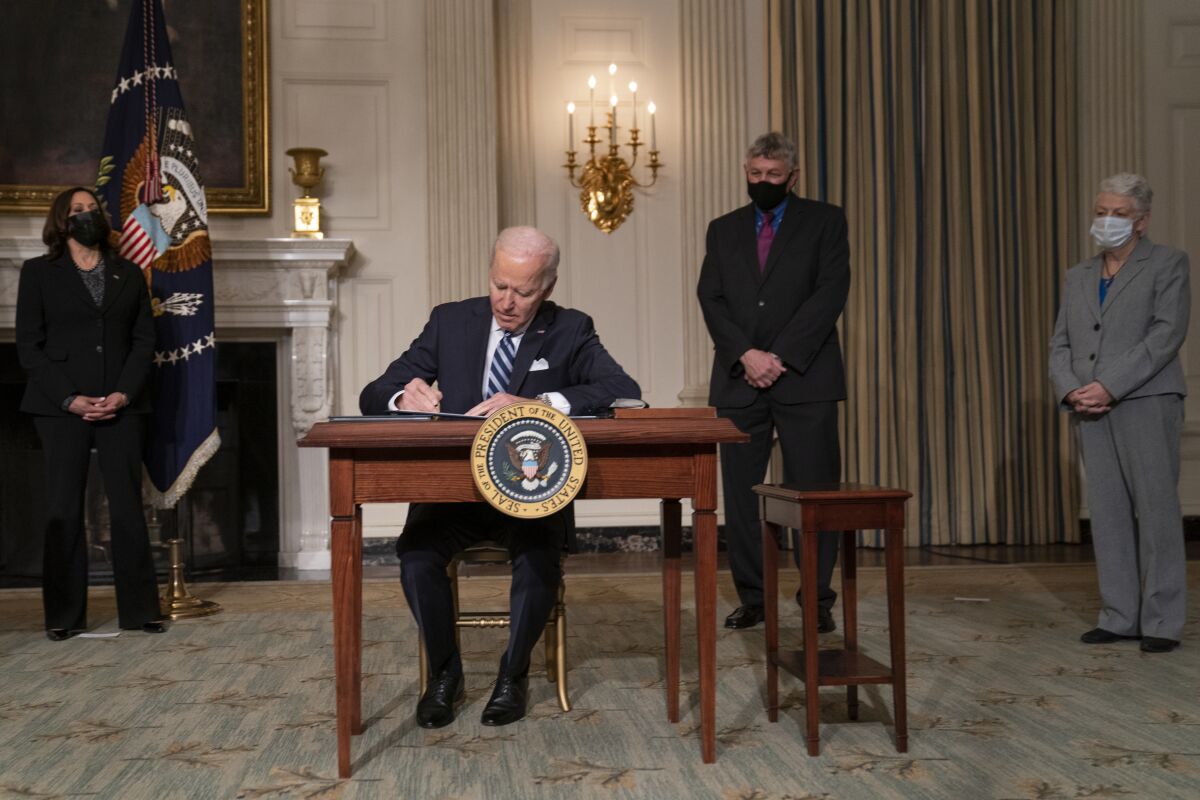 Biden sits at a desk signing documents, with Kamala Harris and two others in the background.