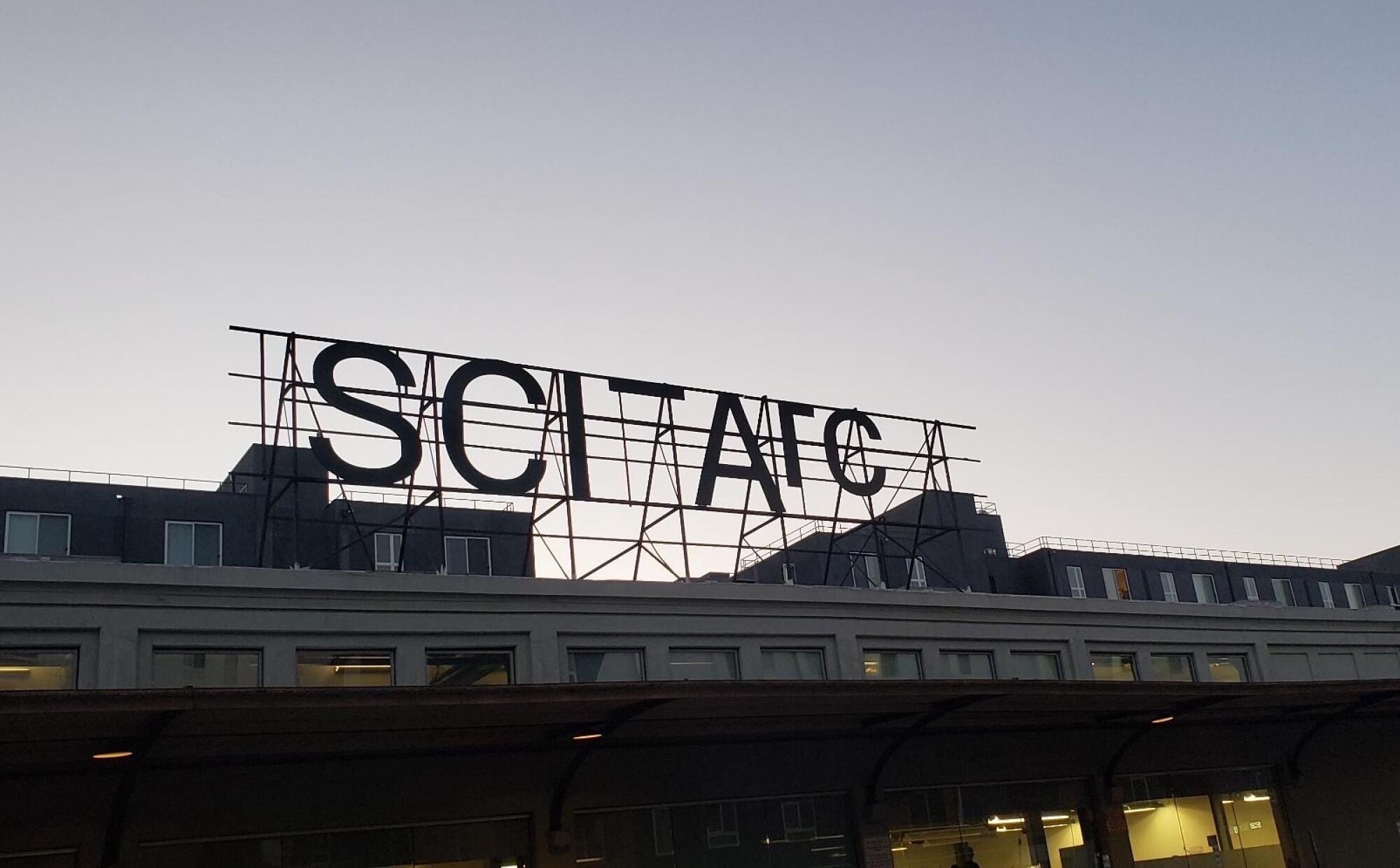 A black metal sign with SCI-Arc's name is silhouetted against a dusky sky