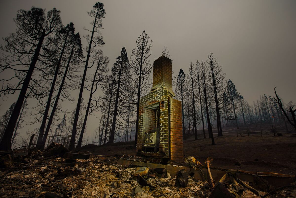 A lone brick chimney stands amid charred ruins of a home under a dark, hazy sky
