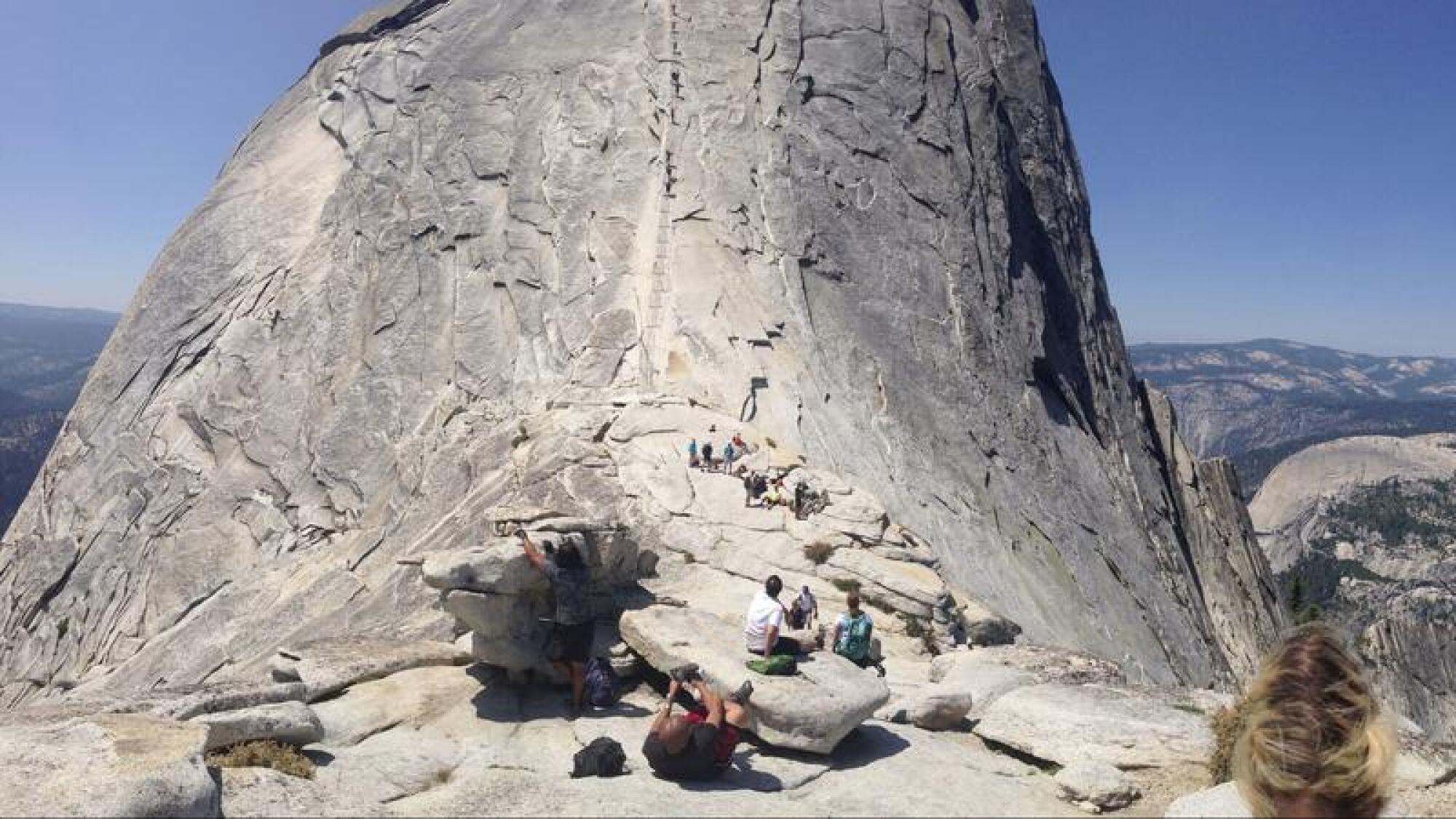 People sit on a boulder in the foreground. In the background hikers ascend bare rockface.