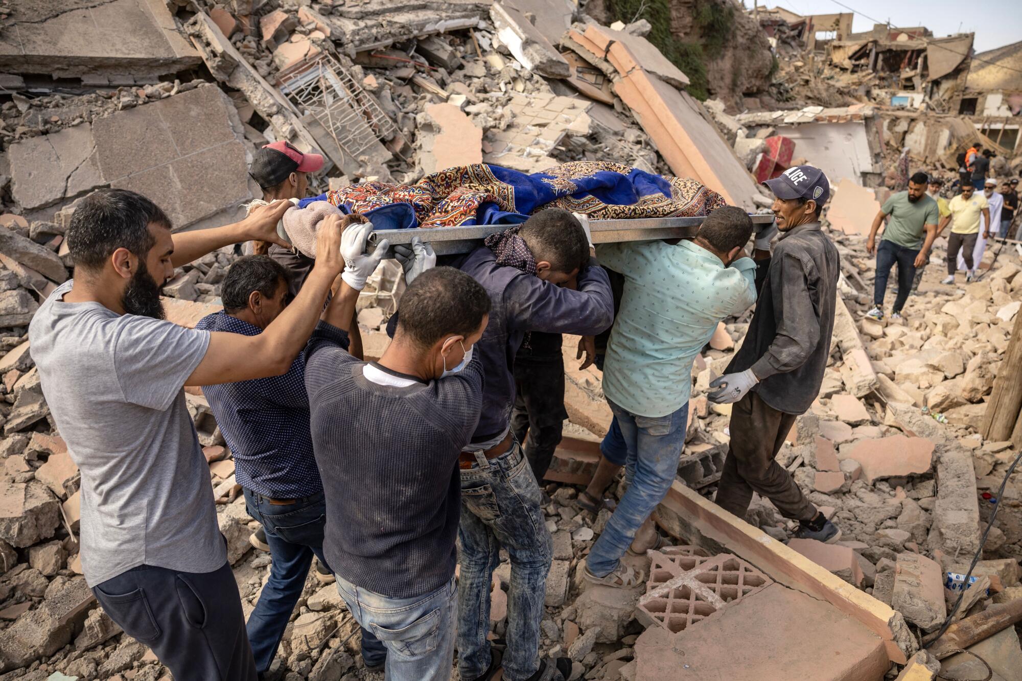 People carry the remains of a victim as they walk over rubble among collapsed buildings.