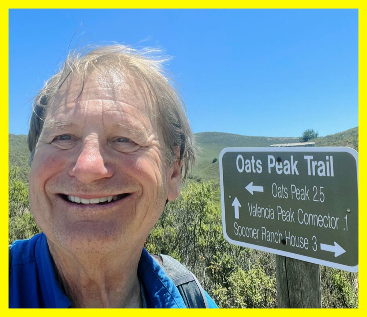 A smiling man outdoors next to a sign for Oats Peak Trail.