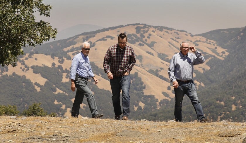 Three men in button-down shirts and jeans walk on an undeveloped site, with steep hills in the background.