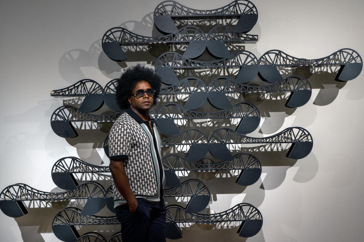 Alexandre Arrechea, a Black man in sunglasses, stands before a wall sculpture resembling a series of conjoined bridges
