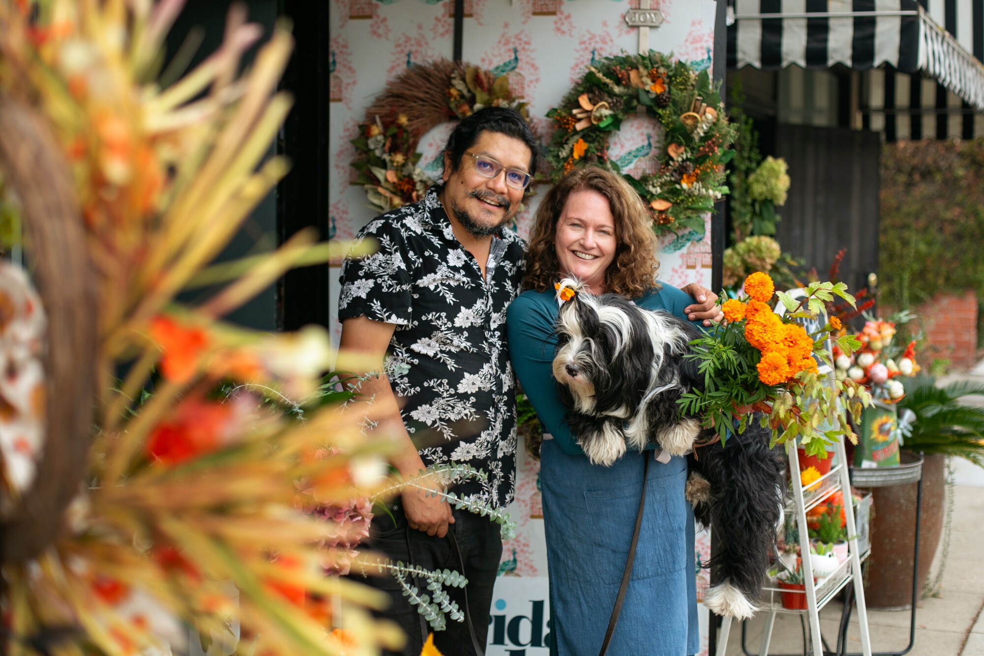 A man and a woman stand in front of their colorful flower shop.  She is holding her black and white dog.