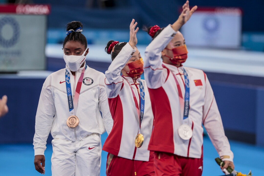 Simone Biles, wearing her medal, walks behind two other gymnasts, who wave to the crowd.