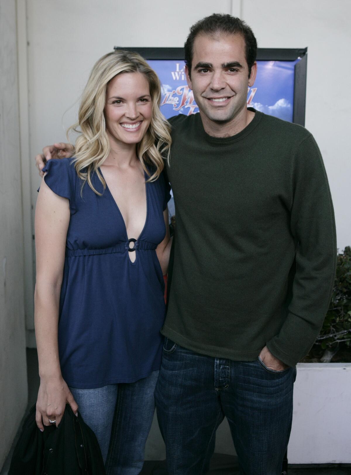 Bridgette Wilson-Sampras smiles in a blue blouse and jeans as she is hugged by Pete Sampras in a green long-sleeve shirt.