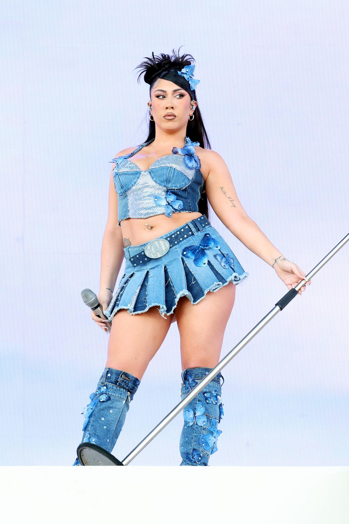 A woman in a blue bustier top, cheerleader skirt and boots tilts a mic stand