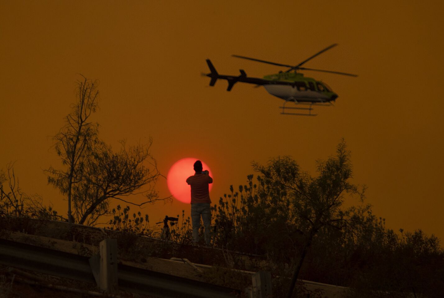 Chris O'Neal, a video journalist from Laguna Niguel, is silhouetted by a smoke-obscured sun as a fire helicopter keeps an eye on the Holy fire approaching McVicker Canyon.