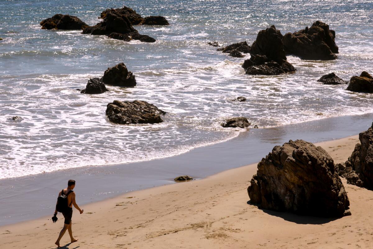 A barefoot person in swim trunks and wearing a backpack walks near boulders on a sandy beach.