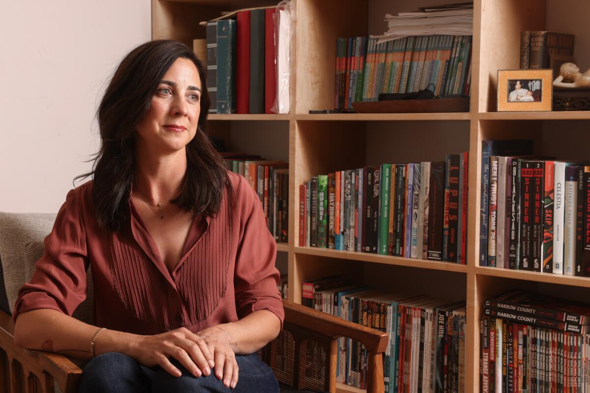 A smiling woman with long dark hair sits in front of a bookshelf full of books.