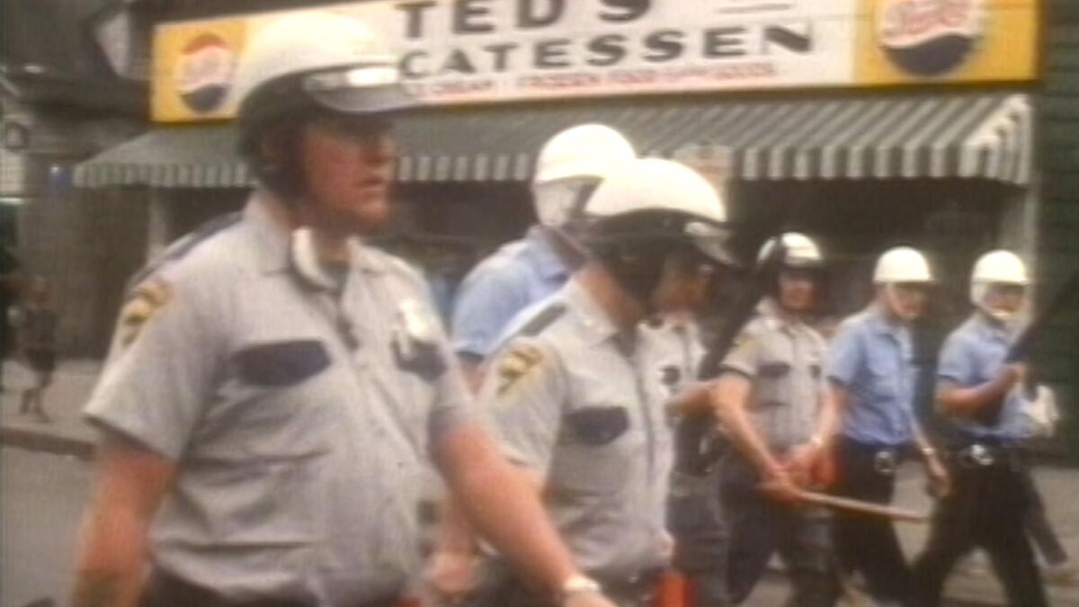 Police officers walk down a street in grainy period video footage.