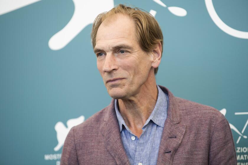 Julian Sands in a brown blazer and a blue suit shirt posing against a turquoise backdrop