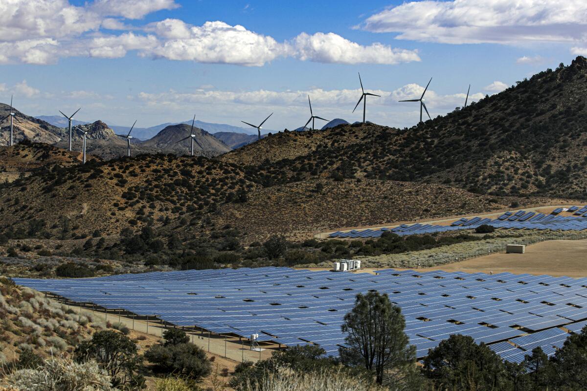 A solar farm in a mountainous setting, with windmills in the background and blue skies with clouds