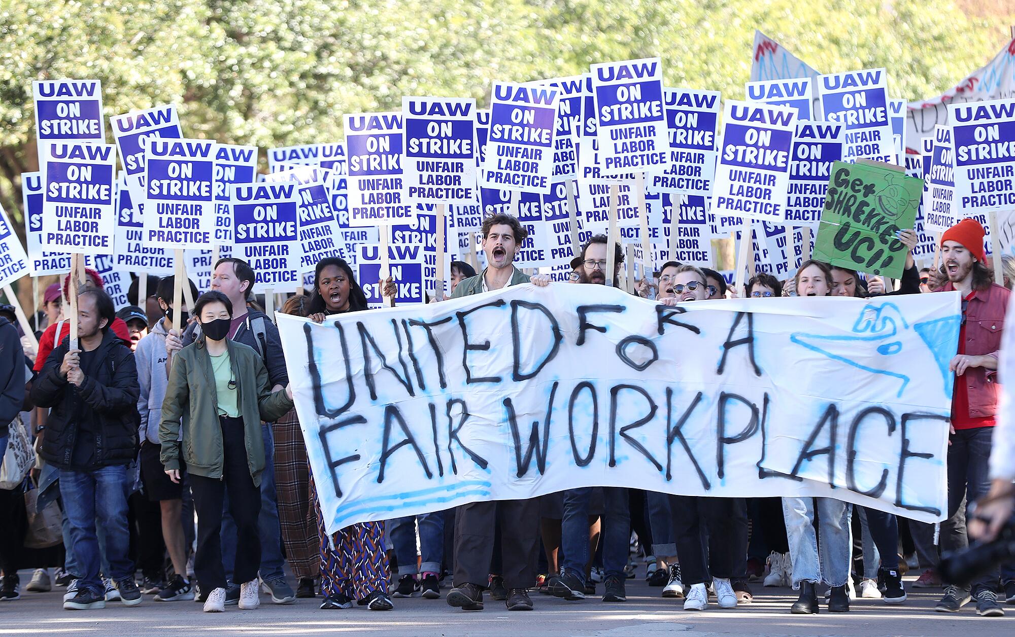 Large group of students walk with signs and a banner that reads "United For a Fair Workplace" at UC Irvine