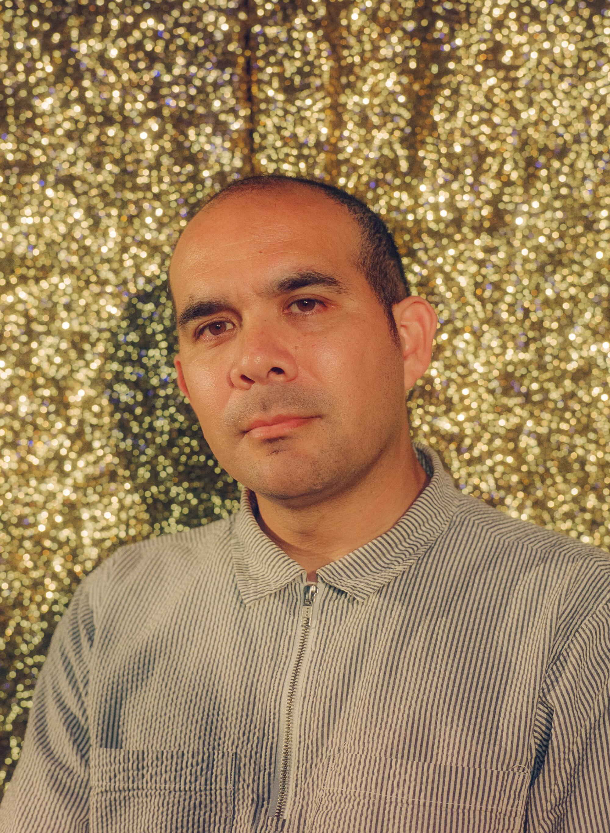 A man in a gray shirt poses in front of a golden curtain.