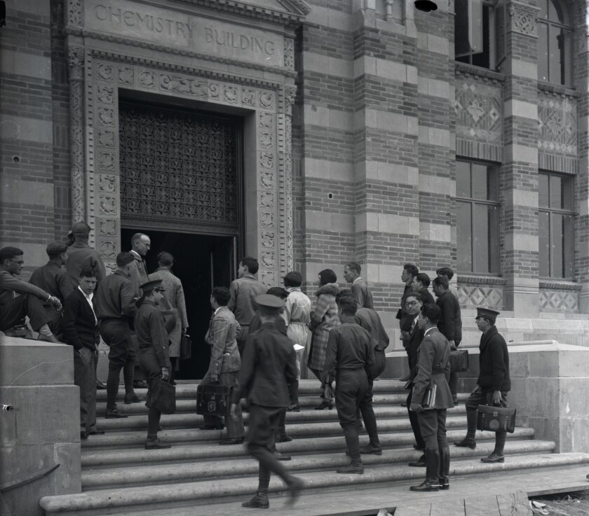 A group of people carrying books and book bags, some wearing hats, walk up the steps to a large brick building.