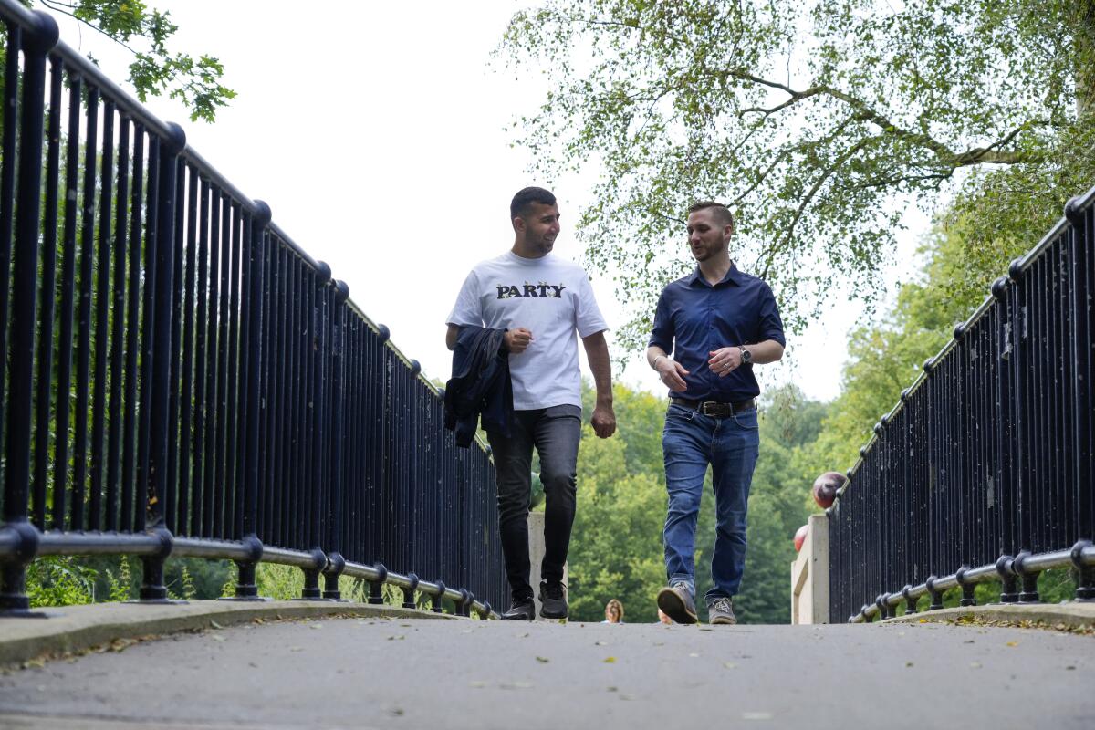 Two men smile as they talk to each other while walking along a fenced path in a park setting 