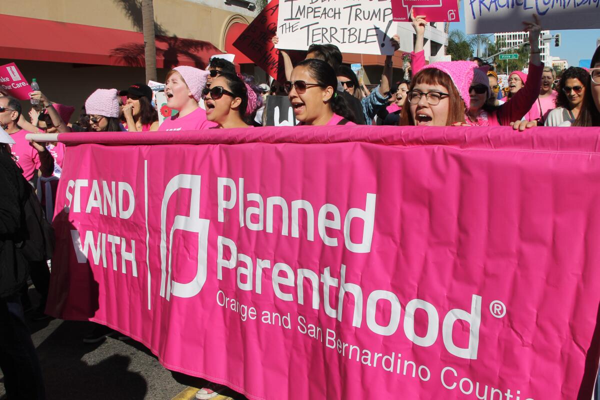 A group of women, many in pink caps, marching and carrying a pink banner reading, "Stand with Planned Parenthood."