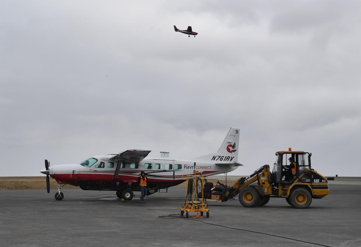 A tractor carrying luggage next to a small plane on an airport tarmac as another plane flies overhead.