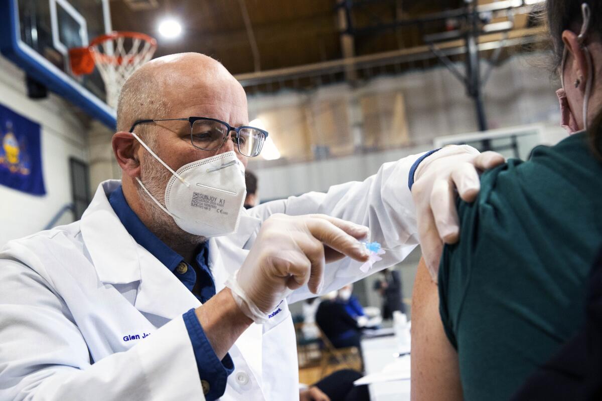 A man in a mask uses a syringe to inject the vaccine into a person's upper arm.