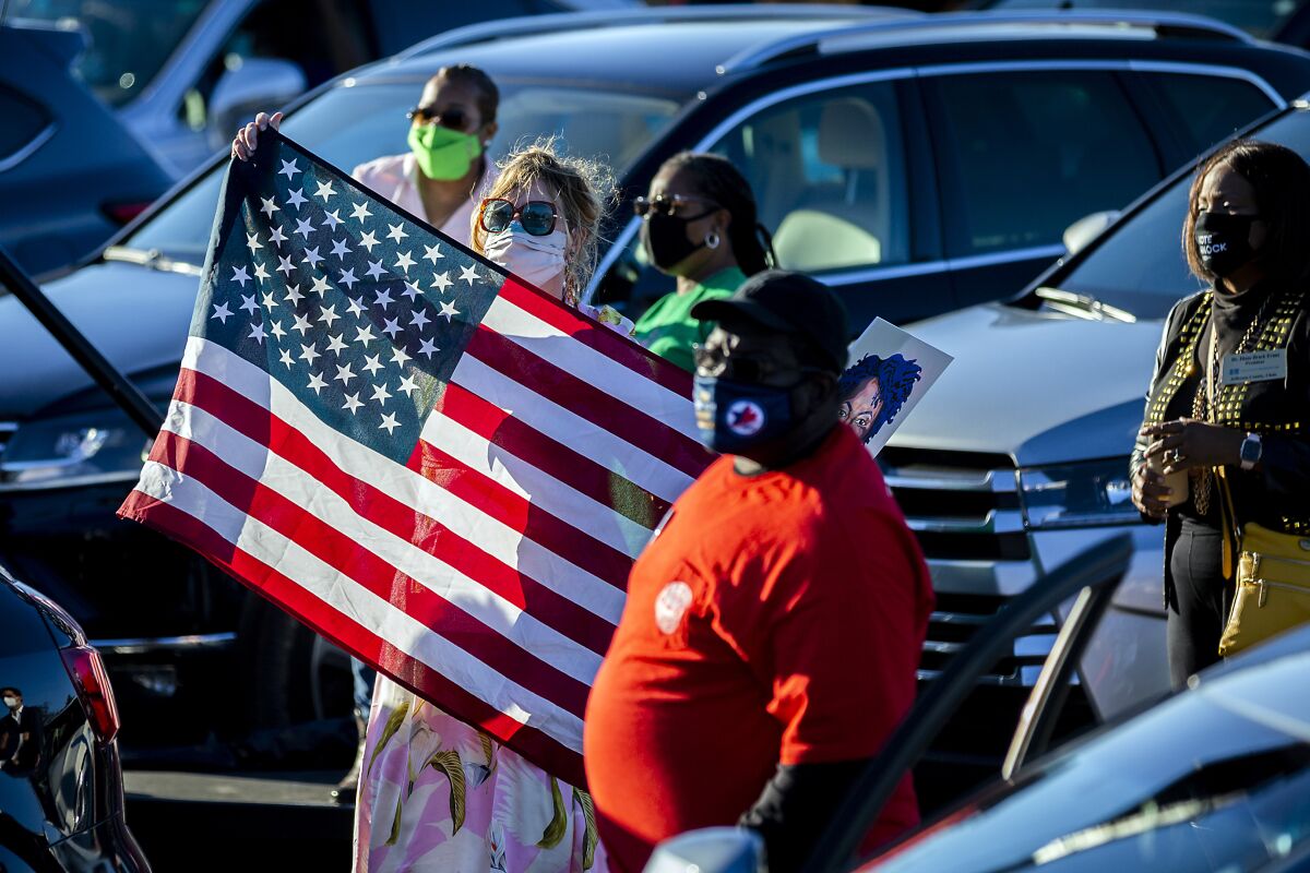 A group of people stand next to cars; one woman holds up a U.S. flag.