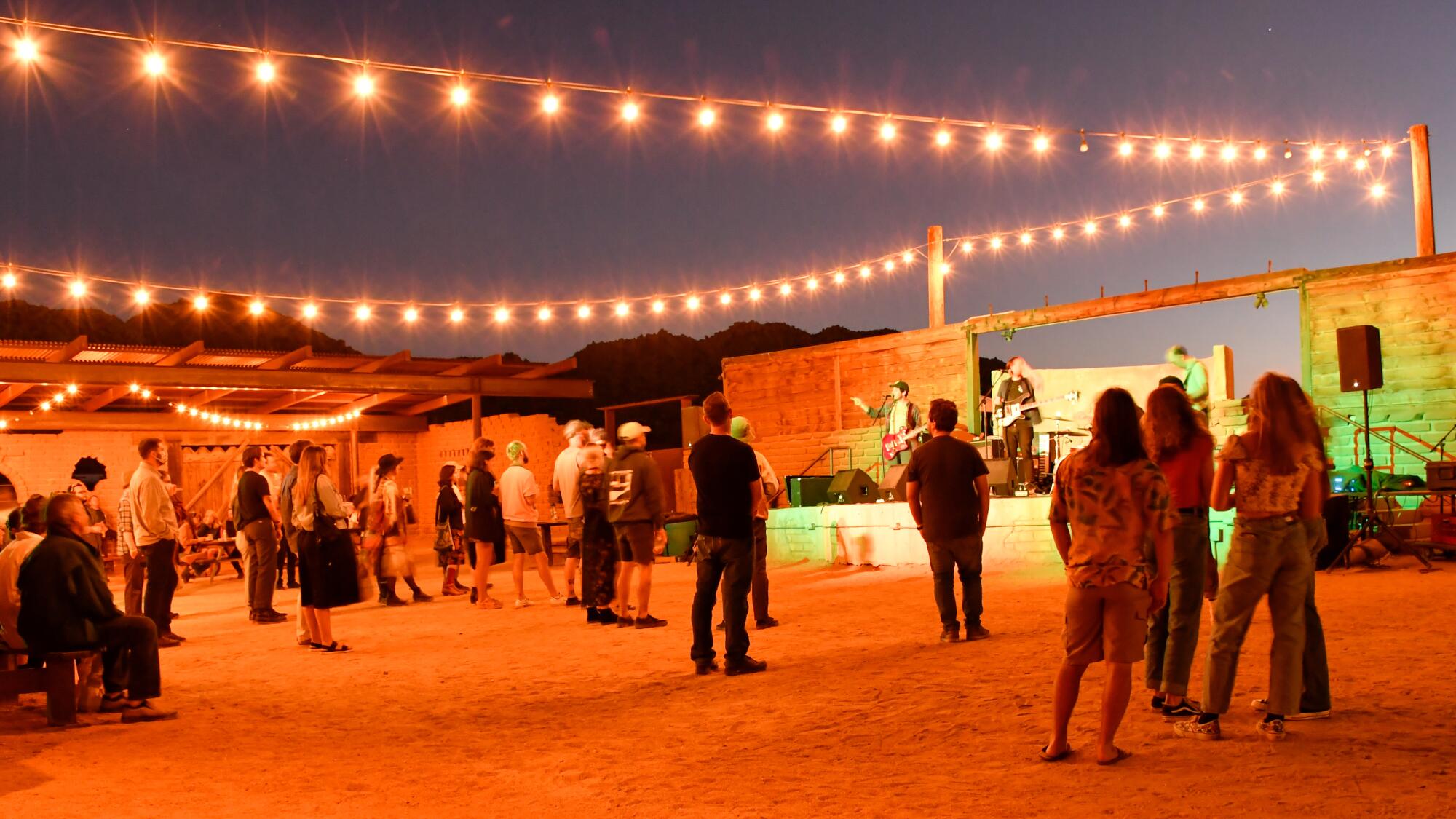 People stand in front of a wood stage on a dirt ground with string lights above 