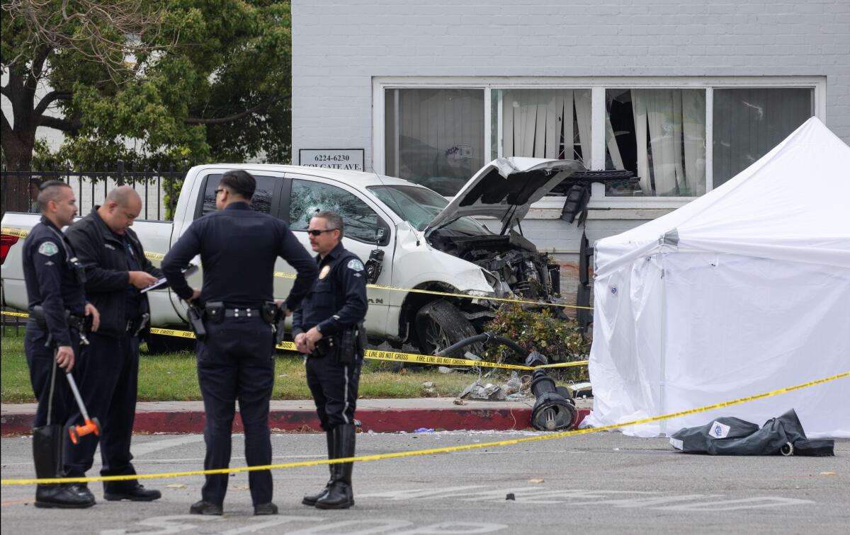 Police officers stand next to a canopy and a crashed pickup truck surrounded by crime scene tape near apartments