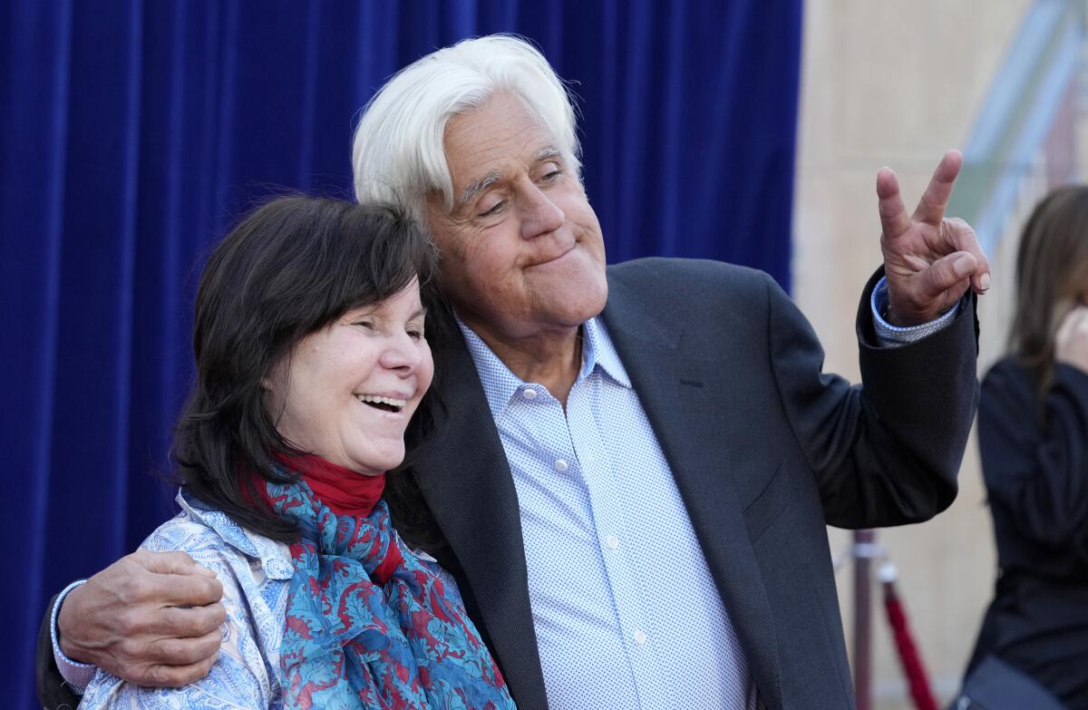 Jay Leno holding one arm around smiling wife Mavis while making a peace sign with his other hand in front of a blue curtain
