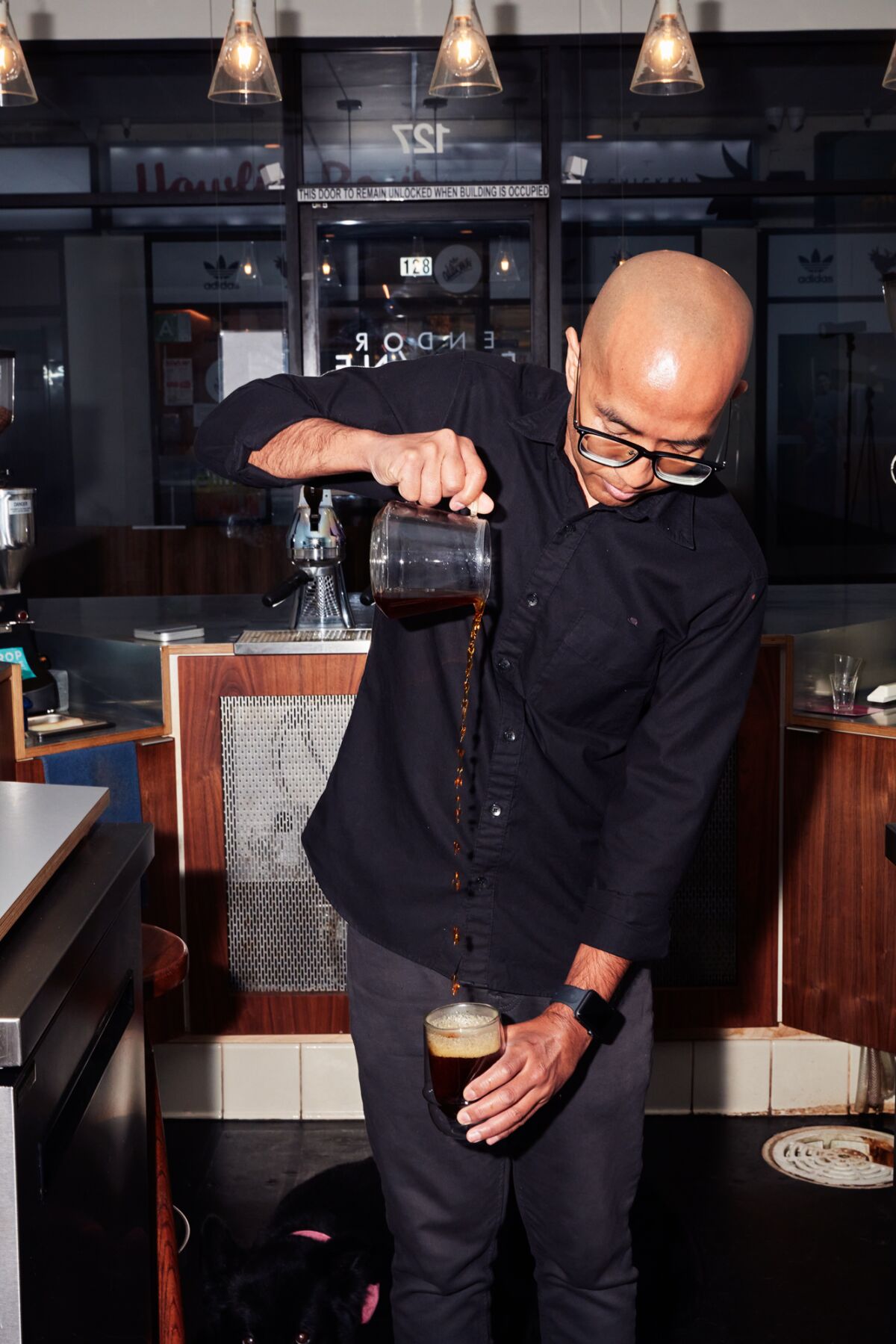 A man raises a hand high to pour into a coffee glass below.