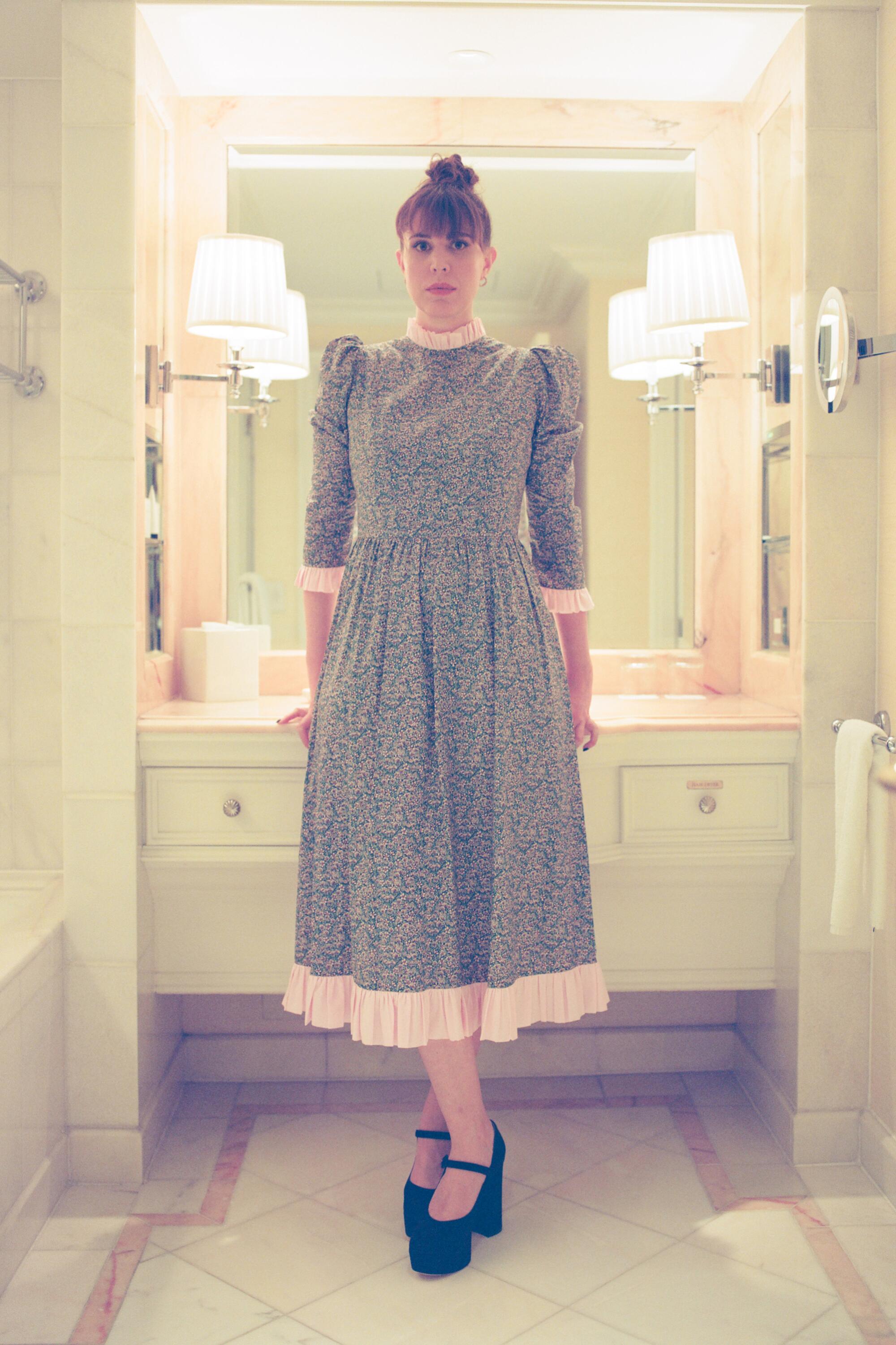 Alice Birch in a modest gray dress and a bun on top of her head stands in a brightly lit hotel bathroom. 