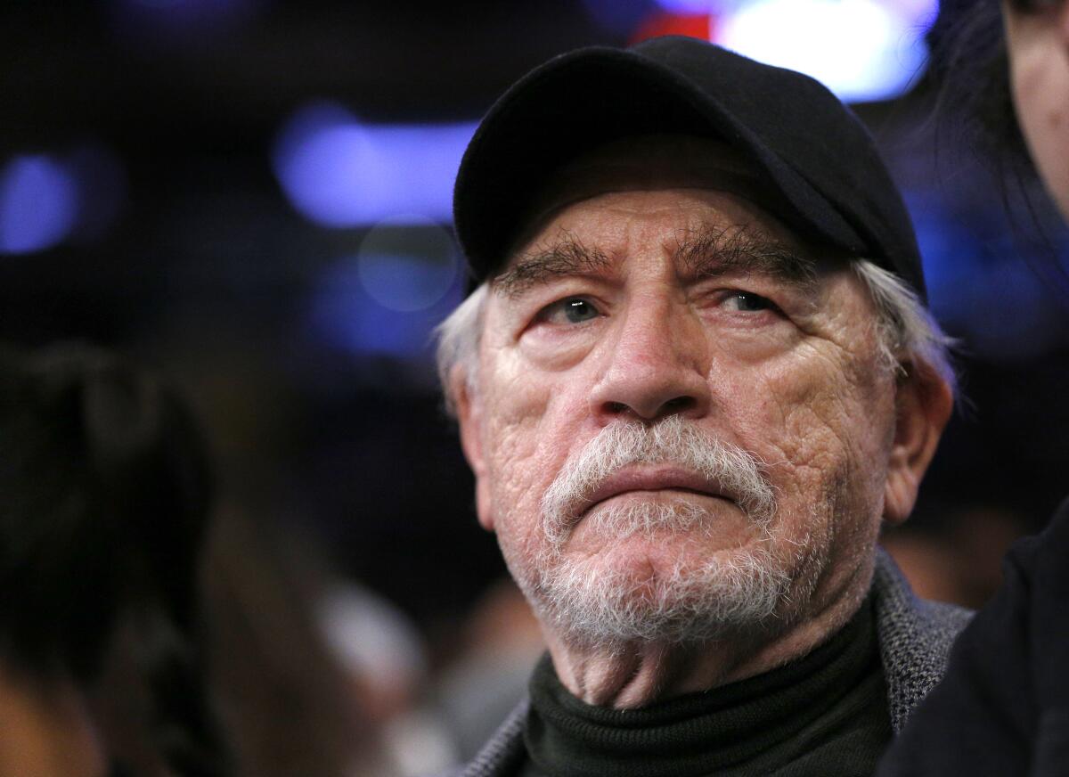 Brian Cox stares past the camera in a black ballcap while attending a sporting event