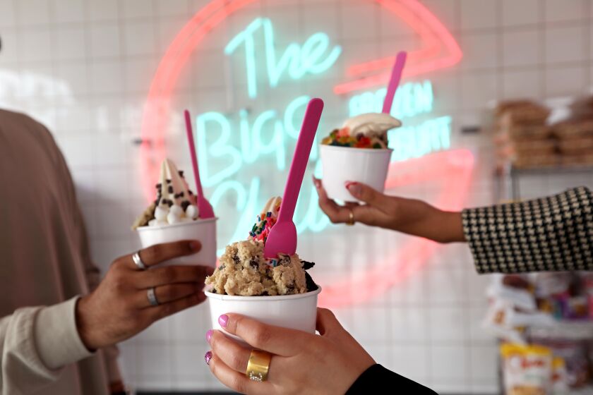Three hands holding cups of frozen yogurt and cookie dough in front of a neon sign