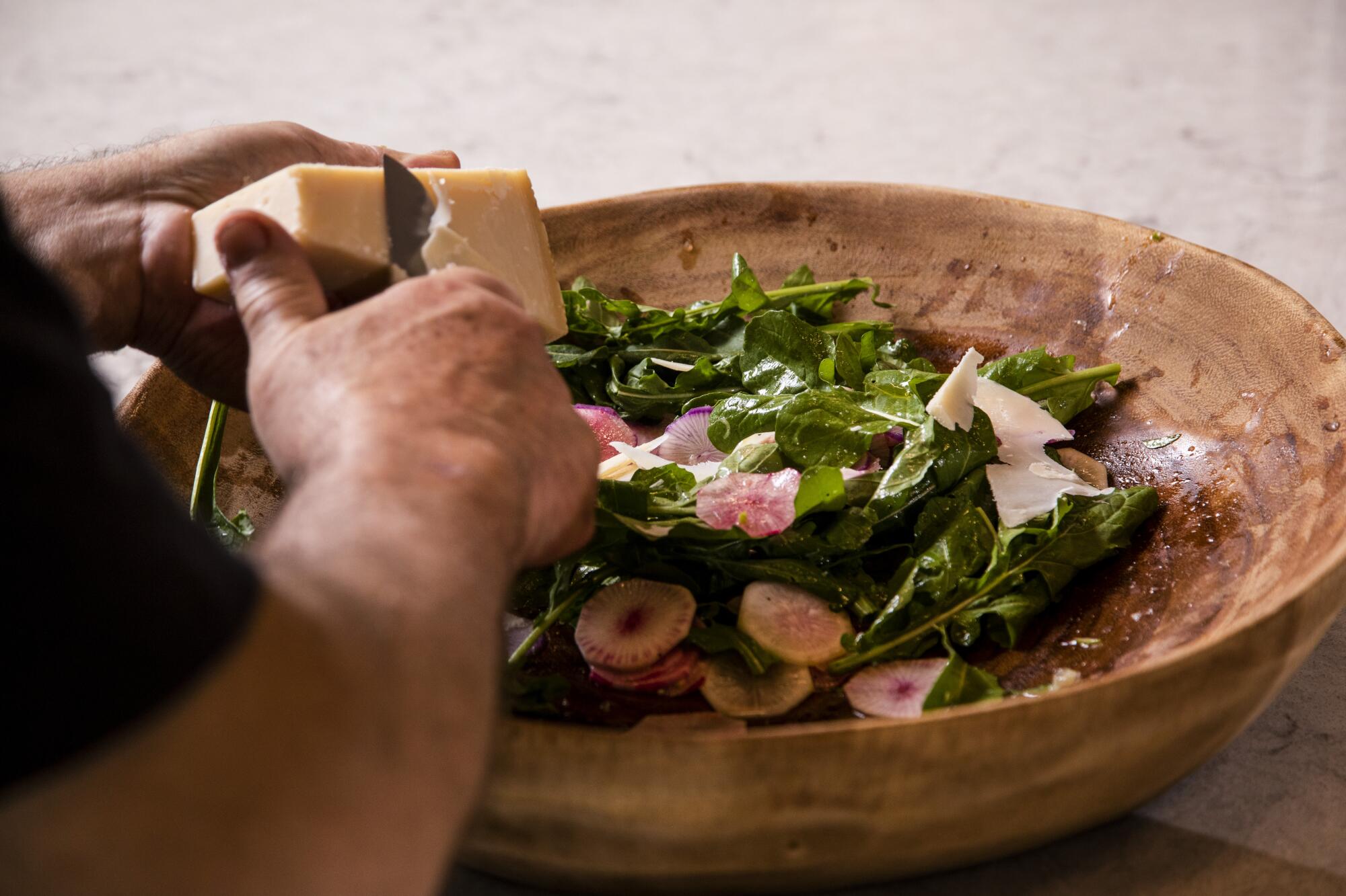 A man slices cheese over a bowl of salad. 