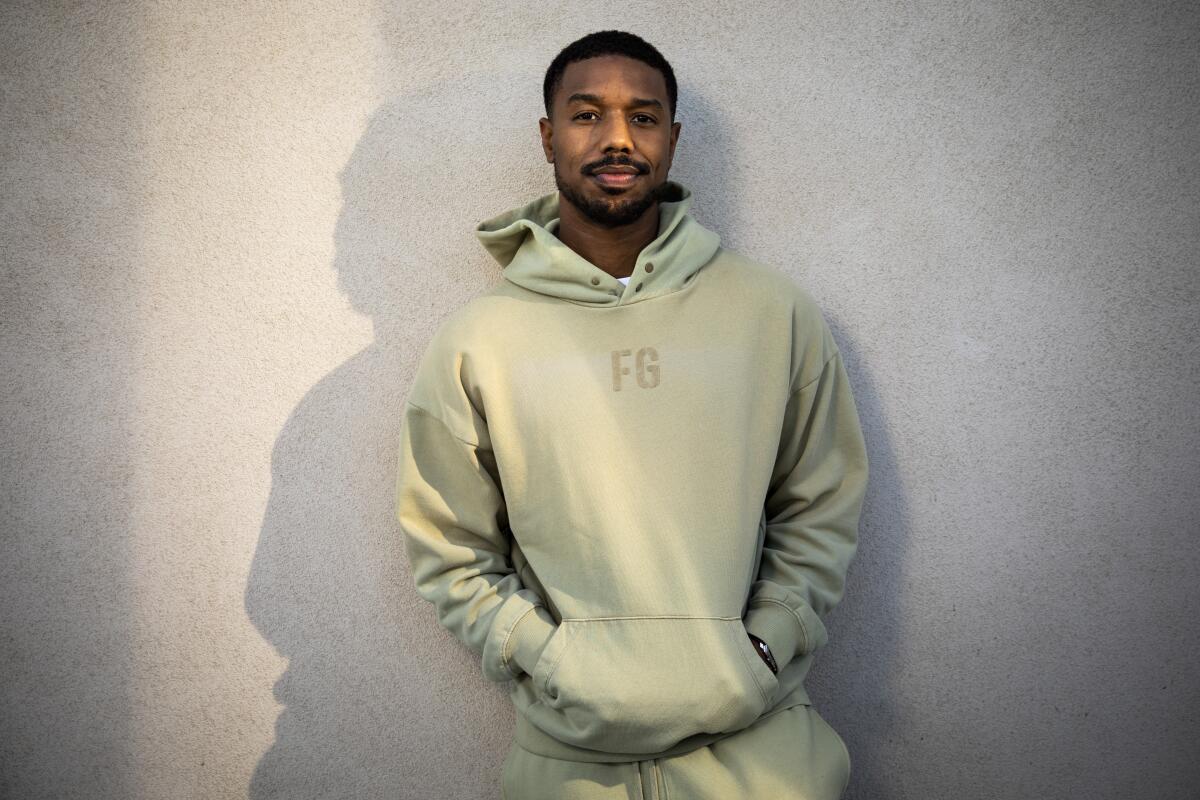 Actor Michael B. Jordan leans against a white wall while wearing a light green hoodie.